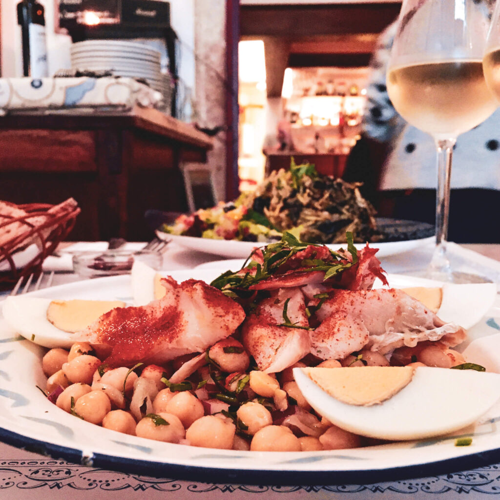 Winteressen in Lissabon: Kichererbsen mit Bacalhau und Ei auf einem Teller, daneben ein Glas Weißwein