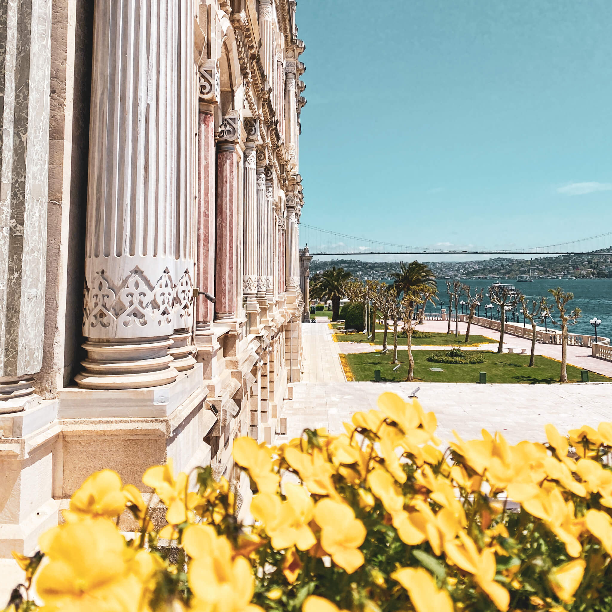 Istanbul, Türkei. Die Fassade des Ciraga-Palastes, davor gelbe Blumen, im Hintergrund der Bosporus mit Brücke.